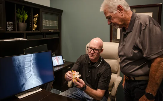 Chiropractor using a spine model to explain spinal anatomy to a patient beside an X-ray display.